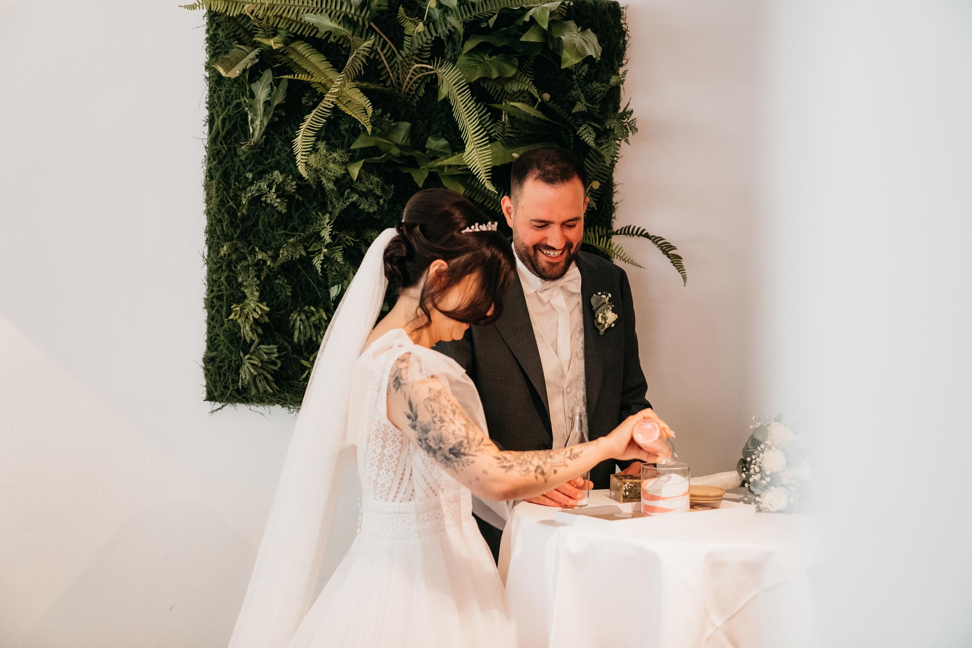 Ein lächelndes Brautpaar bei der Trauzeremonie, mit Pflanzenwand, Brautkleid und floralem Hintergrund.