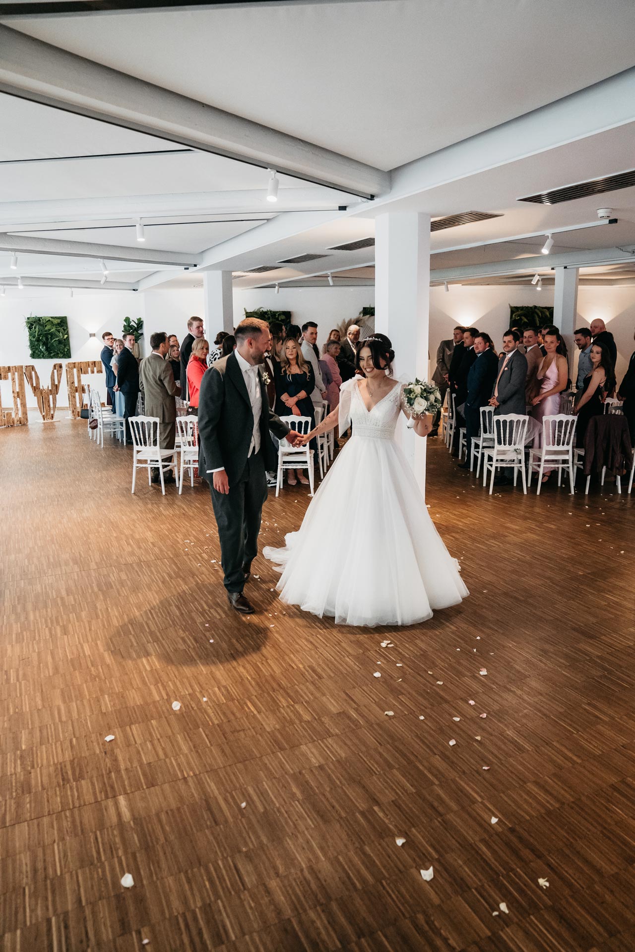 Ein Brautpaar in einem Saal, umgeben von Gästen. Die Braut trägt ein weißes Kleid, der Bräutigam Anzug.