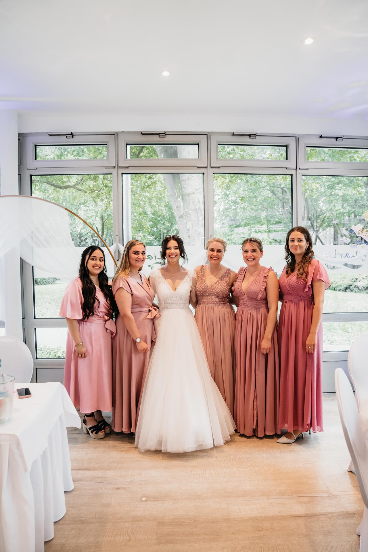 Braut im weißen Kleid mit fünf Brautjungfern in rosa Kleidern lächelnd vor Fenster.