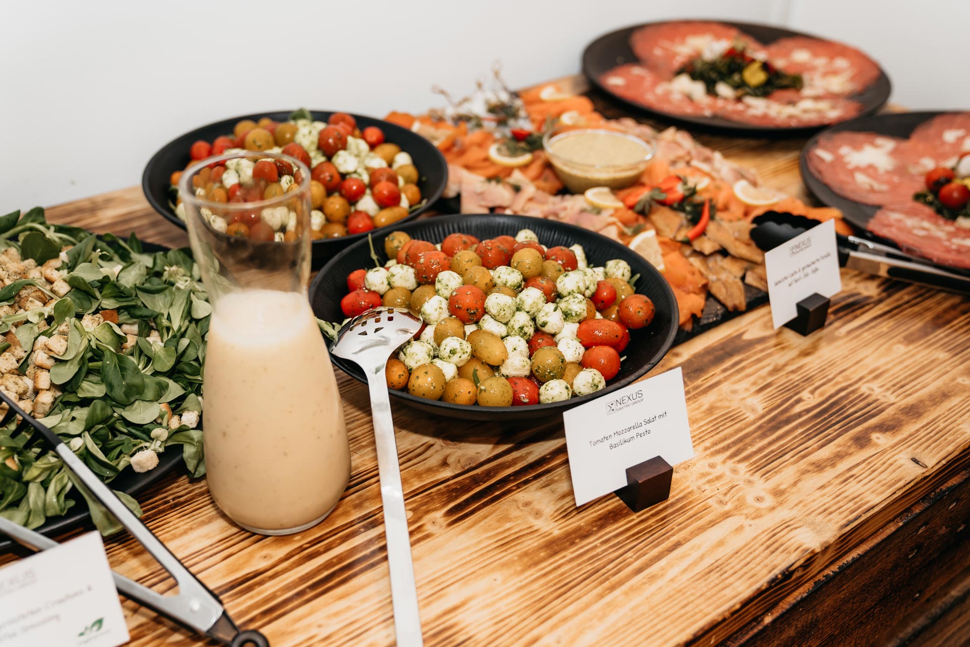 Eine Auswahl an Tellern mit Salaten, Tomaten und Dressing auf einem Holztisch in einem Buffetstil.