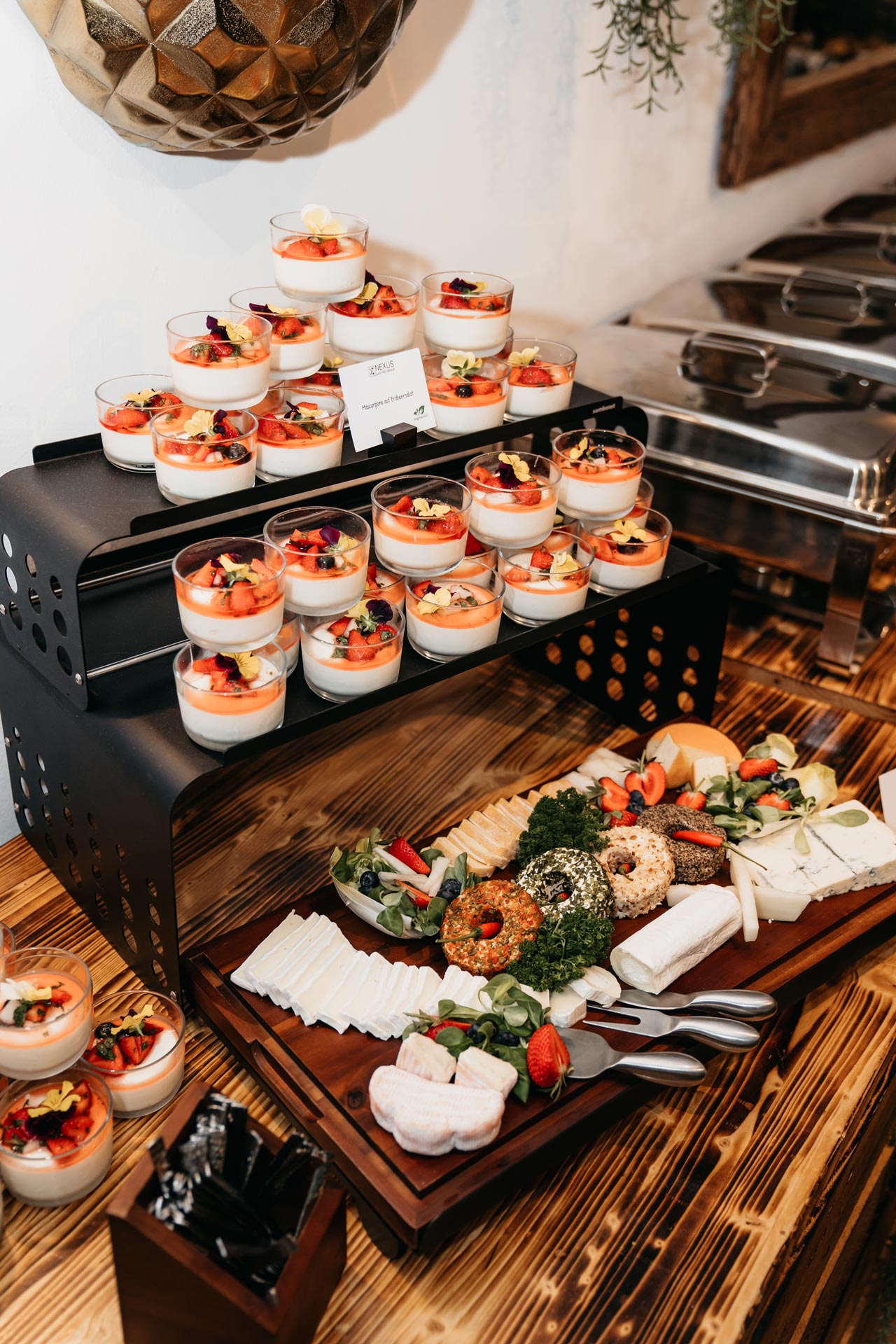 Buffet mit belegten Bagels, geschnittenem Gemüse und Beeren-Desserts in Gläsern, auf Holzplatte.