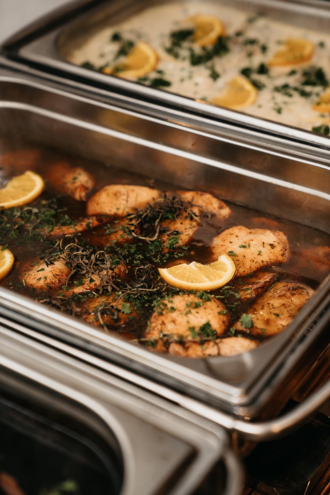 Metallbehälter mit gebratenem Hähnchen, Zitronenscheiben, Kräutern und einem weiteren Gericht im Hintergrund.