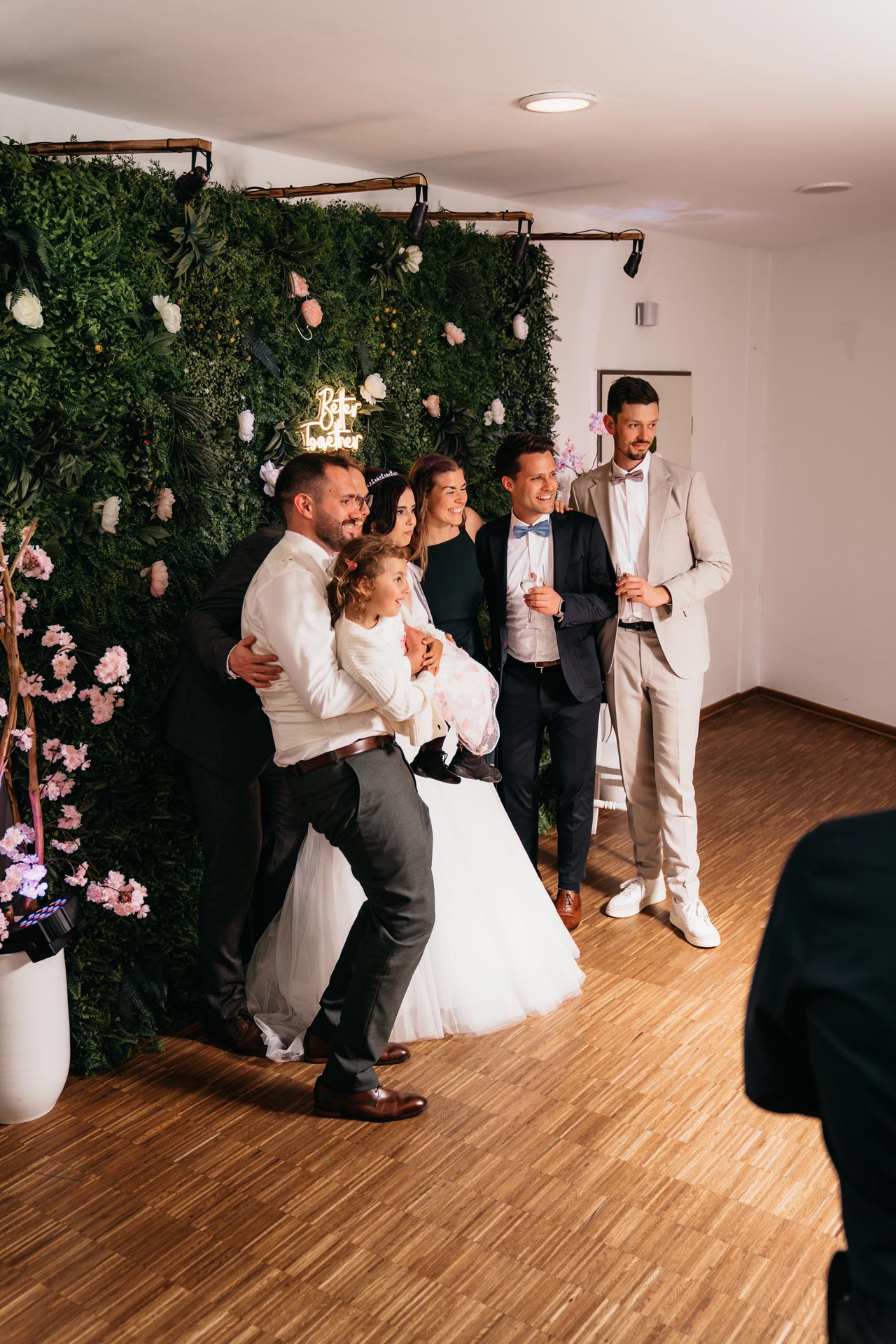 Vier Erwachsene und ein Kind posieren lachend vor einer grünen Blumenwand in eleganter Kleidung.