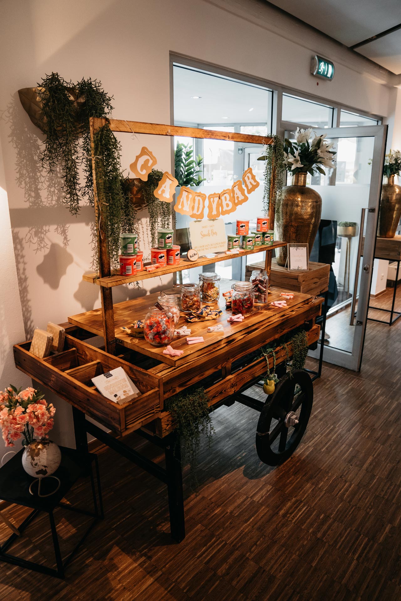 Ein Holzwagen mit Candybar im Raum, dekoriert mit Pflanzen und Süßigkeiten in Gläsern.