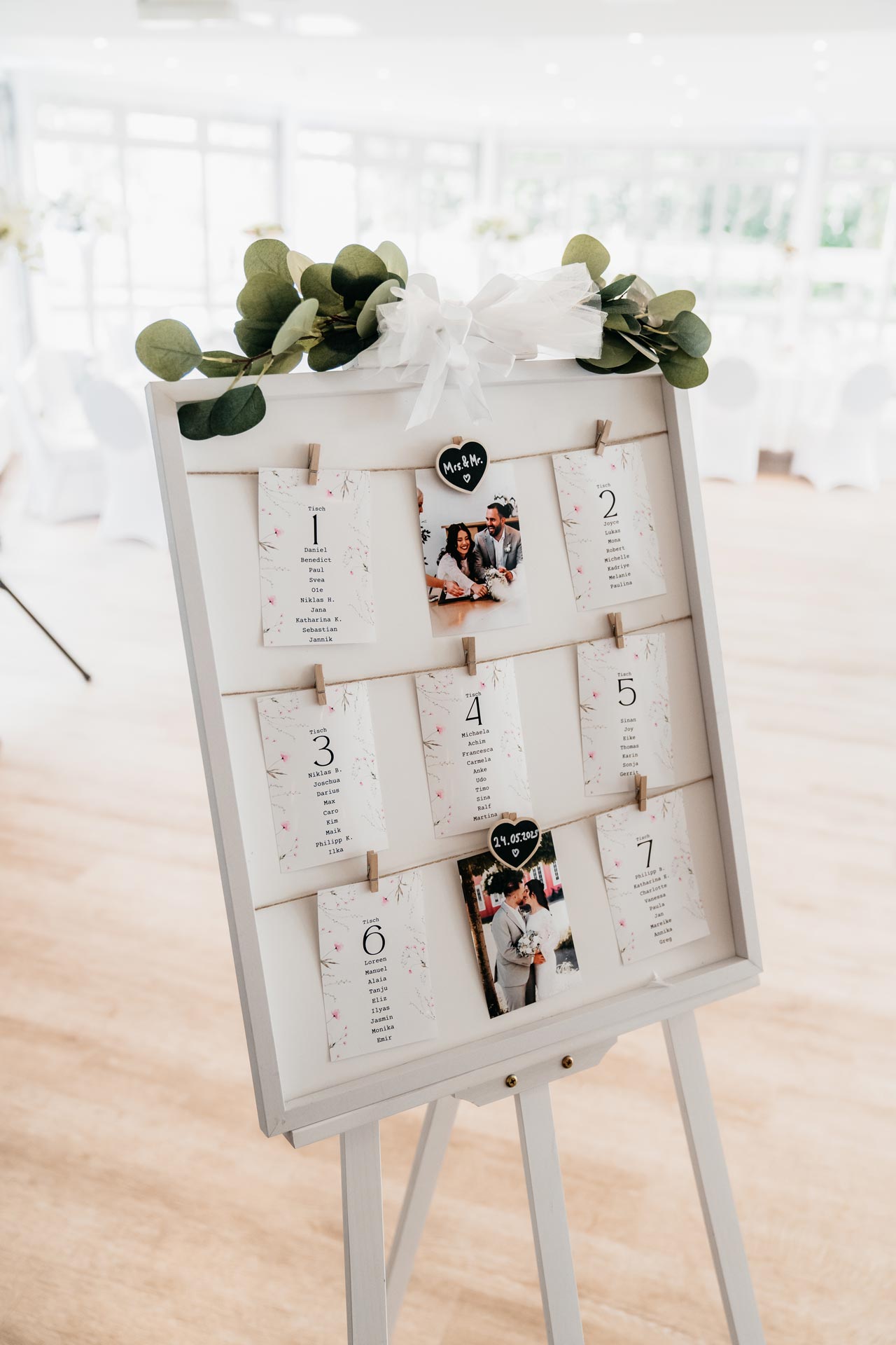 Tafel mit Tischkarten und Fotos, dekoriert mit Eukalyptus und weißer Schleife in hellem Raum.