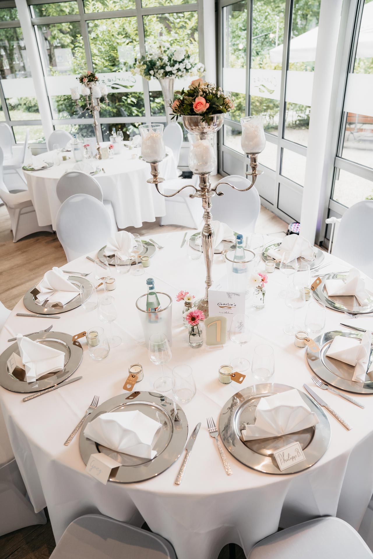 Eleganter Hochzeitstisch mit weißen Servietten, silbernen Akzenten und blumiger Kerzentablette.