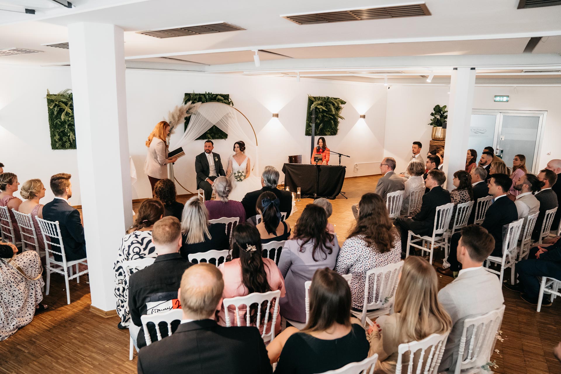 Ein Hochzeitspaar sitzt lächelnd vor einer versammelten Hochzeitsgesellschaft in einem festlichen Saal.