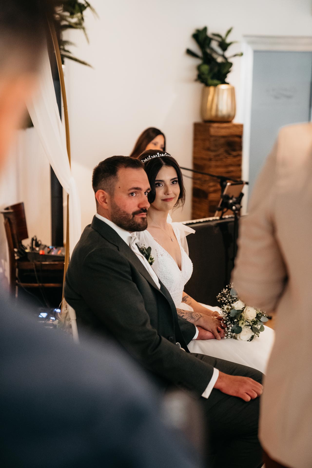 Ein Hochzeitspaar sitzt nebeneinander; sie lächelt, er schaut ernst. Elegantes Setting mit Pflanzen.
