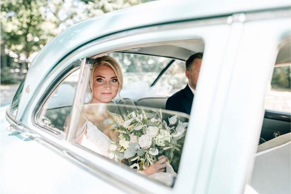 Eine blonde Frau im Brautkleid lächelt freundlich und hält einen Blumenstrauß im Auto