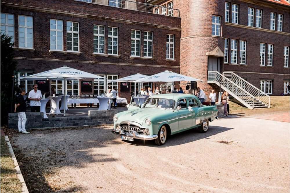 Ein grünes Oldtimer Auto steht vor einem Backsteingebäude mit Menschen im Hintergrund