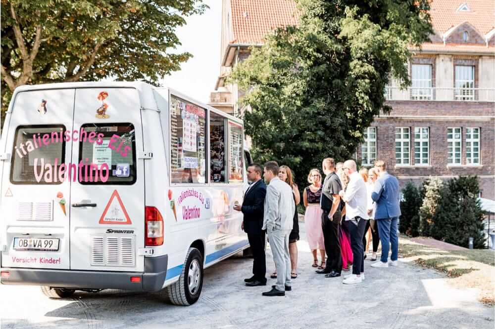 Menschen stehen gut gelaunt an einem weißen Eiswagen mit bunten Aufklebern an