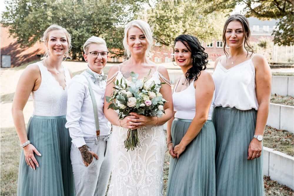 Fünf lächelnde Personen in festlicher Kleidung stehen draußen neben der Braut mit Blumenstrauß