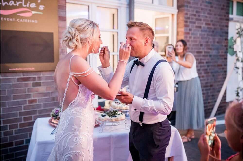 Eine blonde Braut und ein Bräutigam mit Schleife füttern sich lächelnd Kuchen