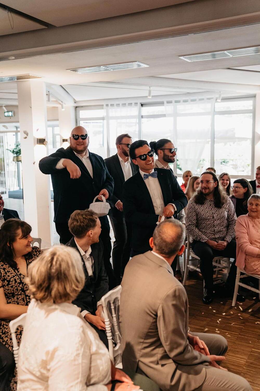 Fünf Männer in Anzügen und Sonnenbrillen lachen locker und stehen unter Gästen